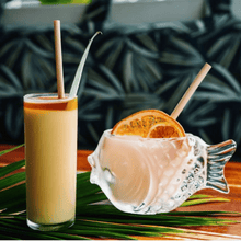 Load image into Gallery viewer, A tall glass of frozen rum drink with a bamboo straw beside a unique fish-shaped glass filled with a creamy pink cocktail, garnished with a dried orange slice, both on a palm leaf against a tropical patterned backdrop
