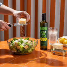 Load image into Gallery viewer, Person grating cheese over a salad with Hassy avocado oil bottle on a tiled surface.