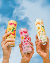 Load image into Gallery viewer, alldae superfruit soda's variety pack held in the air by hands against a blue sky background, as a nod to the gentle afternoon boost these provide.
