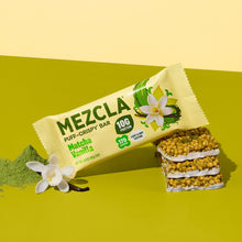 Load image into Gallery viewer, Mezcla Matcha Vanilla puff-crispy protein bar package on green surface with vanilla flowers and matcha powder, showing stacked bar pieces with white cream layers.