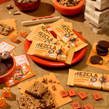 Load image into Gallery viewer, MEZCLA peanut butter chocolate protein bars arranged on orange plate surrounded by games, nuts, and snacks on wooden surface