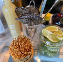 Load image into Gallery viewer, Eco-friendly straws from Holy City Straw Co. in a wooden holder, placed next to cocktail-making tools, lime wedges in a jar, and a bottle of mixer on a bar counter.