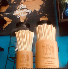 Load image into Gallery viewer, Wooden holders filled with eco-friendly straws from Holy City Straw Co., displayed on a counter with glass jars and a 'Big Travelling Man' sign in the background.