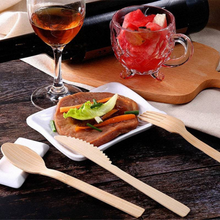 Load image into Gallery viewer, Eco-friendly bamboo cutlery from Holy City Straw Co. displayed on a wooden table next to a square white plate with a grilled steak garnished with a lettuce leaf and carrot strips. A glass of amber-colored drink and a cup of fruit salad complete the cozy dining setting.
