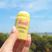 Load image into Gallery viewer, Hand holding a yellow sunscreen stick with a blue sky and greenery in the background
