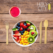 Load image into Gallery viewer, Top view of a wooden table setting featuring Holy City Straw Co. brand bamboo cutlery, with a bamboo fork on the left and a knife on the right, framing a colorful bowl of fresh fruit salad containing strawberries, blueberries, kiwi, and mango cubes. Above the bowl is a glass of red smoothie, and the Holy City Straw Co. logo with a wheat stalk icon is prominently displayed at the top.
