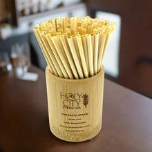 Load image into Gallery viewer, A cylindrical bamboo holder branded with ‘HolyCity Straw Co. – The Earth Straw,’ filled with a large bundle of natural bamboo straws. The holder’s etched text mentions gluten-free, biodegradable features, and it rests on a blurred tabletop background.
